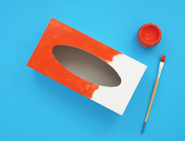 Une boîte de mouchoirs vide, de la peinture rouge et un pinceau pour fabriquer un instrument fait maison.