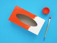 Une boîte de mouchoirs vide, de la peinture rouge et un pinceau pour fabriquer un instrument fait maison.