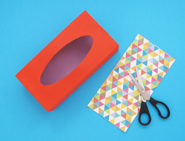 Boîte de mouchoirs vide peinte en rouge avec une paire de ciseaux et un morceau rectangulaire de papier décoratif