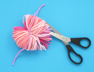 Une paire de ciseaux noirs coupe le bout des boucles de laine pour obtenir des fils, toujours avec la ficelle de laine centrale rose qui maintient le tout