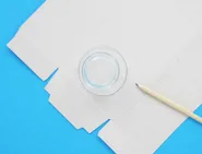 Une boîte de mouchoirs en carton est ouverte et mise à plat sur une surface bleue avec un petit verre et un crayon