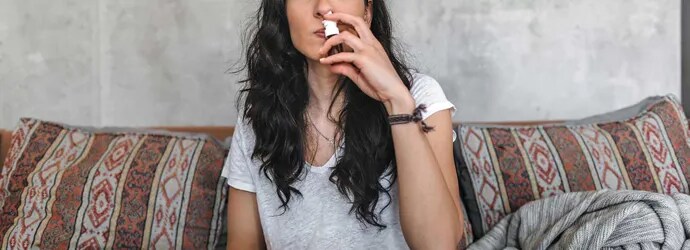 Une jeune femme avec une allergie au pollen de bouleau est assise sur le canapé et porte un spray nasal à son nez