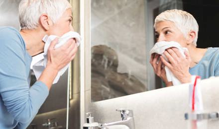 Une femme avec des  cheveux blancs et courts regarde dans le miroir de salle de bain après avoir rincé son visage pour soulager ses yeux larmoyants à cause de l'allergies