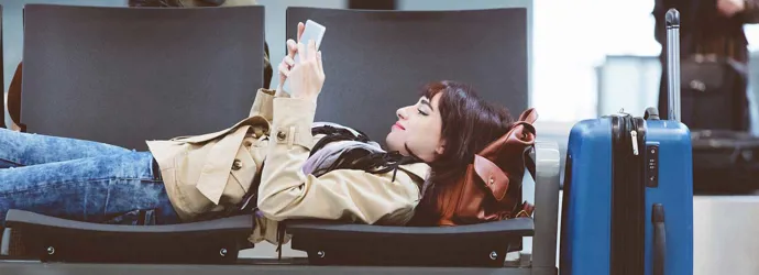 Une femme installée sur plusieurs sièges dans un aéroport lit un livre avec une valise à roulettes à côté d'elle