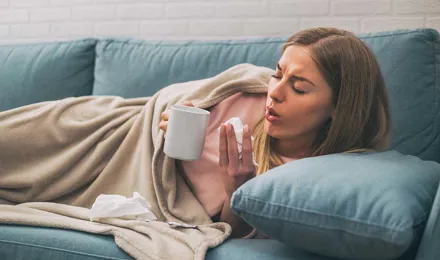 Une femme tousse couchée dans canapé tout en tenant une tasse de thé et un mouchoir