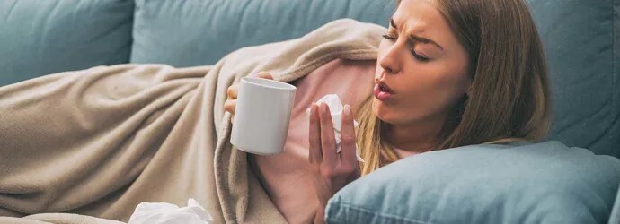 Une femme tousse couchée dans canapé tout en tenant une tasse de thé et un mouchoir