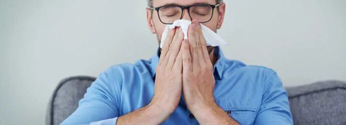 Un homme qui porte des lunettes et une chemise bleue est assis sur un canapé et se mouche le nez