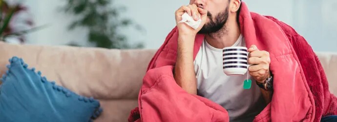 Nuori mies yskii sohvalla peiton alla kädessään teemuki ja nenäliinoja